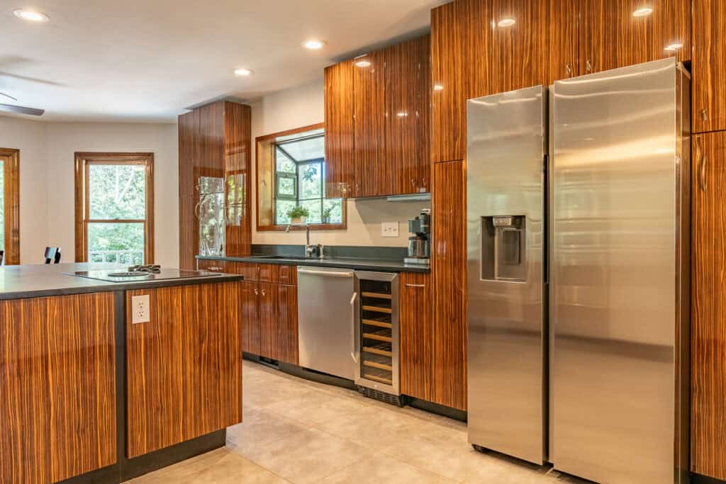 A sleek kitchen with stainless steel refrigerator, wine cooler, and wood-paneled cabinets, illuminated by recessed lighting.