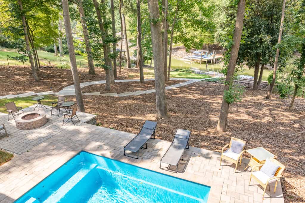 An elevated view of a backyard featuring a bright blue pool, patio seating, and a wooded area leading to a lake dock.