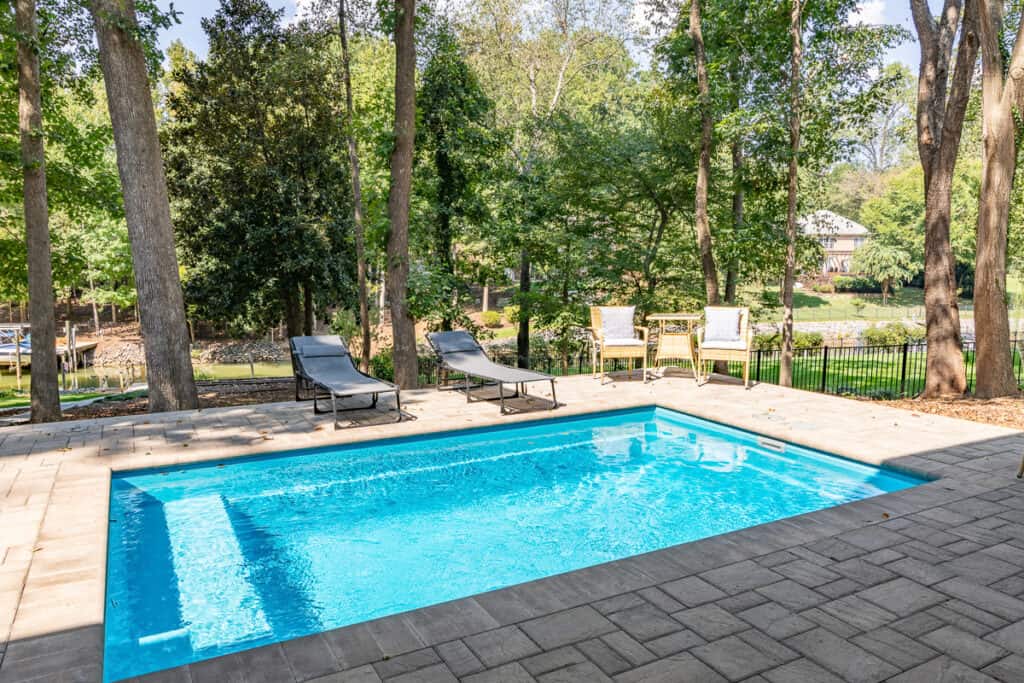 A tranquil pool area with sun loungers and chairs, set against a backdrop of tall trees and a distant view of a neighboring property.