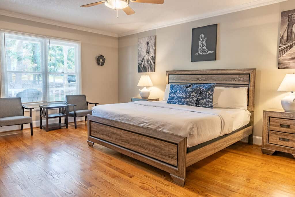 A spacious master bedroom with a king-sized bed, hardwood floors, and a cozy sitting area near a large window offering natural light.